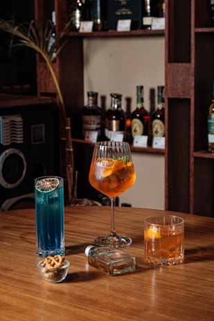 Three different types of cocktails in distinct glasses are placed on a wooden tabletop. Behind them, bottles of alcohol are arranged neatly on wooden shelves against the wall. There is a blue cocktail with a slice of dried lime, an orange cocktail in a large goblet glass with slices of orange and other fruits, and a shorter glass with another orange drink containing citrus slices. Additionally, a small glass bowl with a few pretzels and a clear glass bottle also sit on the table.