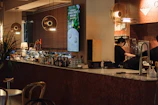 A cozy, modern bar with a sleek marble countertop and various bottles of alcohol displayed behind it. On the left side, there are several hanging round lights with a warm glow. A large digital screen is mounted on the wall displaying an advertisement for a Single Orzo Pancake. Two people are working behind the counter, one pouring a drink. To the left, a low table and chair setup can be seen along with a potted plant adding natural elements to the space.