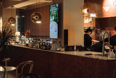 A cozy, modern bar with a sleek marble countertop and various bottles of alcohol displayed behind it. On the left side, there are several hanging round lights with a warm glow. A large digital screen is mounted on the wall displaying an advertisement for a Single Orzo Pancake. Two people are working behind the counter, one pouring a drink. To the left, a low table and chair setup can be seen along with a potted plant adding natural elements to the space.