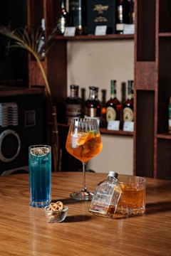 A selection of alcoholic beverages arranged on a wooden table. There are three drinks: a tall blue cocktail, an orange-tinted drink in a wine glass with slices of orange, and a whiskey glass with a small labeled bottle next to it. A small bowl of mixed nuts is also present. Shelves in the background display various bottles of spirits.