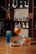 A selection of alcoholic beverages arranged on a wooden table. There are three drinks: a tall blue cocktail, an orange-tinted drink in a wine glass with slices of orange, and a whiskey glass with a small labeled bottle next to it. A small bowl of mixed nuts is also present. Shelves in the background display various bottles of spirits.
