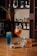A selection of alcoholic beverages arranged on a wooden table. There are three drinks: a tall blue cocktail, an orange-tinted drink in a wine glass with slices of orange, and a whiskey glass with a small labeled bottle next to it. A small bowl of mixed nuts is also present. Shelves in the background display various bottles of spirits.