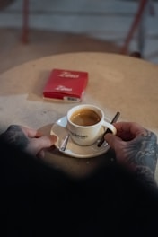 A tattooed hand holds a white cup filled with espresso on a saucer, with a small spoon placed on it. A cigar or cigarette is held in the same hand. In the background, a pack of Zino cigars is slightly out of focus on a round table with a textured surface.