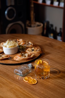 A wooden table is set with an assortment of food and drinks. A glass of orange cocktail is garnished with slices of dried orange, and beside it lies a small, clear bottle. On a round wooden platter are several pieces of bruschetta topped with shrimp and olives. A small white bowl contains potato chips, and next to it is a dish of olives. The background features shelves lined with bottles.
