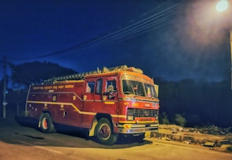 A red fire truck is parked on a dimly lit roadside at night. It has detailed yellow and white markings, including text in a language that seems to be Kannada. The scene is illuminated by a streetlight, reflecting off the truck's surface. The surrounding environment includes a dark sky and silhouetted trees.