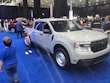 A white pickup truck is displayed in an indoor auto show setting, surrounded by a blue carpet. Several people, including adults and children, are interacting with the vehicle. Another red vehicle is visible in the background, and there are banners and lights hanging from the ceiling.