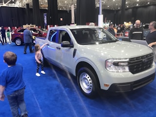 A white pickup truck is displayed in an indoor auto show setting, surrounded by a blue carpet. Several people, including adults and children, are interacting with the vehicle. Another red vehicle is visible in the background, and there are banners and lights hanging from the ceiling.