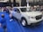 A white pickup truck is displayed in an indoor auto show setting, surrounded by a blue carpet. Several people, including adults and children, are interacting with the vehicle. Another red vehicle is visible in the background, and there are banners and lights hanging from the ceiling.