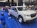 A white pickup truck is displayed in an indoor auto show setting, surrounded by a blue carpet. Several people, including adults and children, are interacting with the vehicle. Another red vehicle is visible in the background, and there are banners and lights hanging from the ceiling.