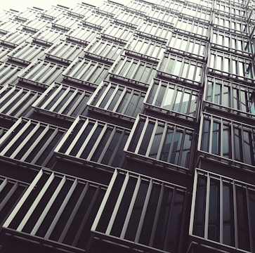 A geometric facade of a modern building with repetitive metal and glass window panels creates a pattern. The architectural design features sleek lines and symmetry, combining dark reflective glass with metallic accents.