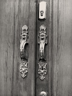 An intricately carved double door features ornate metallic handles and a keyhole set against a wooden background. The design reflects vintage aesthetics with elaborate patterns on the handles and locks.