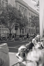 A group of runners is seen participating in a race on a city street, with spectators gathered along the side watching. The scene is in black and white, featuring a large building with classical architecture in the background and trees lining the sidewalk.