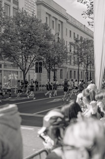 A group of runners is seen participating in a race on a city street, with spectators gathered along the side watching. The scene is in black and white, featuring a large building with classical architecture in the background and trees lining the sidewalk.