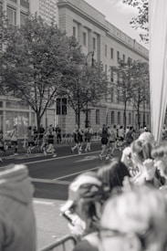A group of runners is seen participating in a race on a city street, with spectators gathered along the side watching. The scene is in black and white, featuring a large building with classical architecture in the background and trees lining the sidewalk.