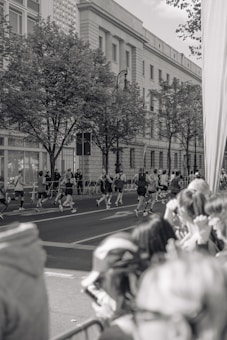 A group of runners is seen participating in a race on a city street, with spectators gathered along the side watching. The scene is in black and white, featuring a large building with classical architecture in the background and trees lining the sidewalk.