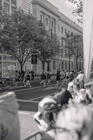 A group of runners is seen participating in a race on a city street, with spectators gathered along the side watching. The scene is in black and white, featuring a large building with classical architecture in the background and trees lining the sidewalk.