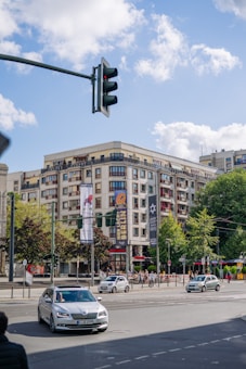 An urban street scene featuring a large building with multiple stories and balconies. The facade has signage with the word 'Berlin.' Several cars are on the road, and pedestrians are walking on the sidewalks. A traffic light is visible against a blue sky with scattered clouds.