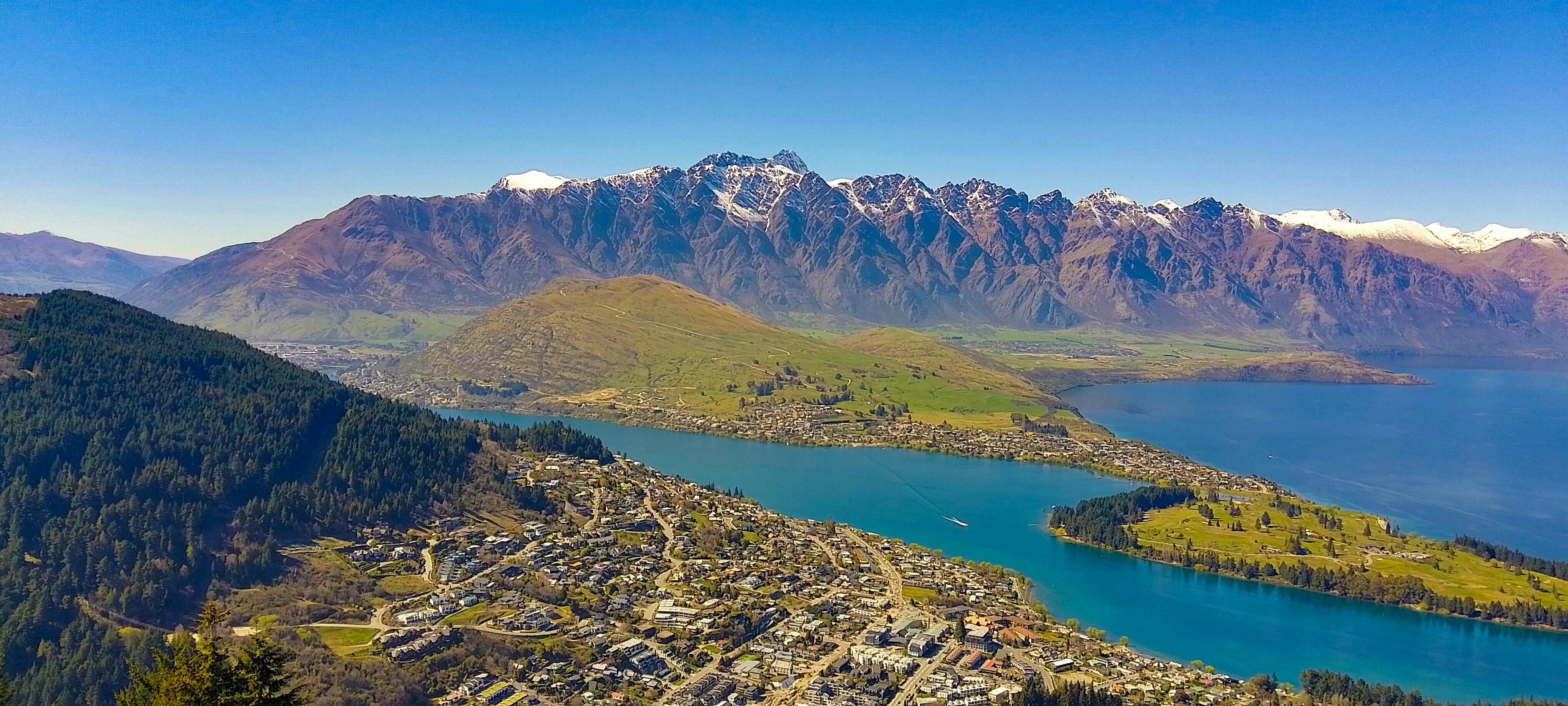 A panoramic view of a picturesque lakeside town nestled at the foot of rugged, snow-capped mountains. The vibrant blue lake is prominent, with boats visible on the water, and the settlement spreads out into the green and brown patchwork landscape leading to the towering peaks under a clear blue sky.