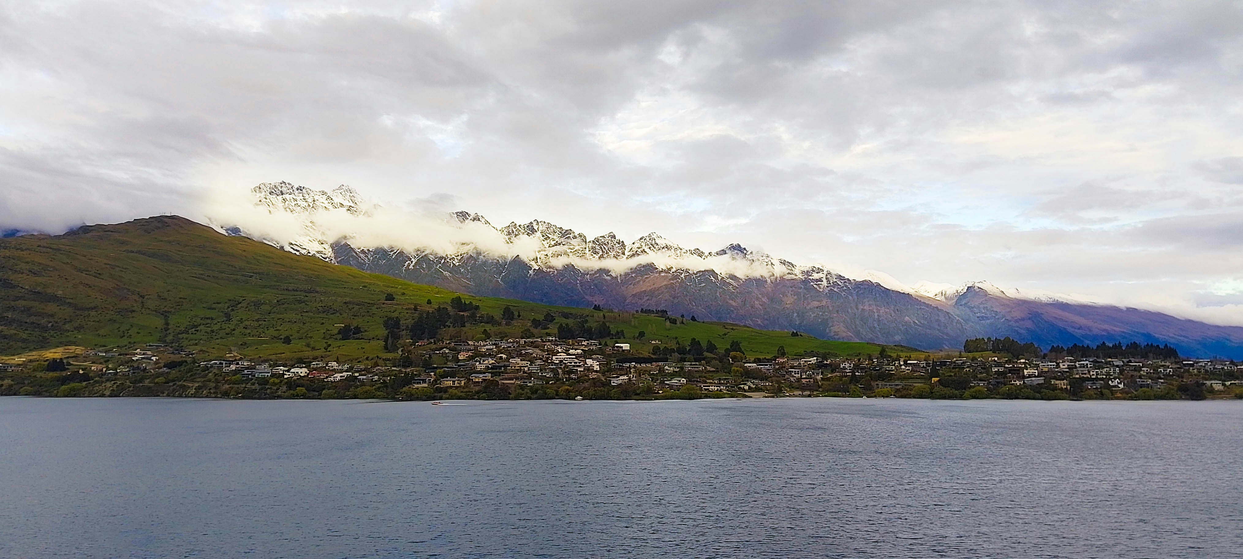 A wide shot of Queenstown across a lake. The sky is cloudy, and behind the town are mountains with snow-capped peaks.