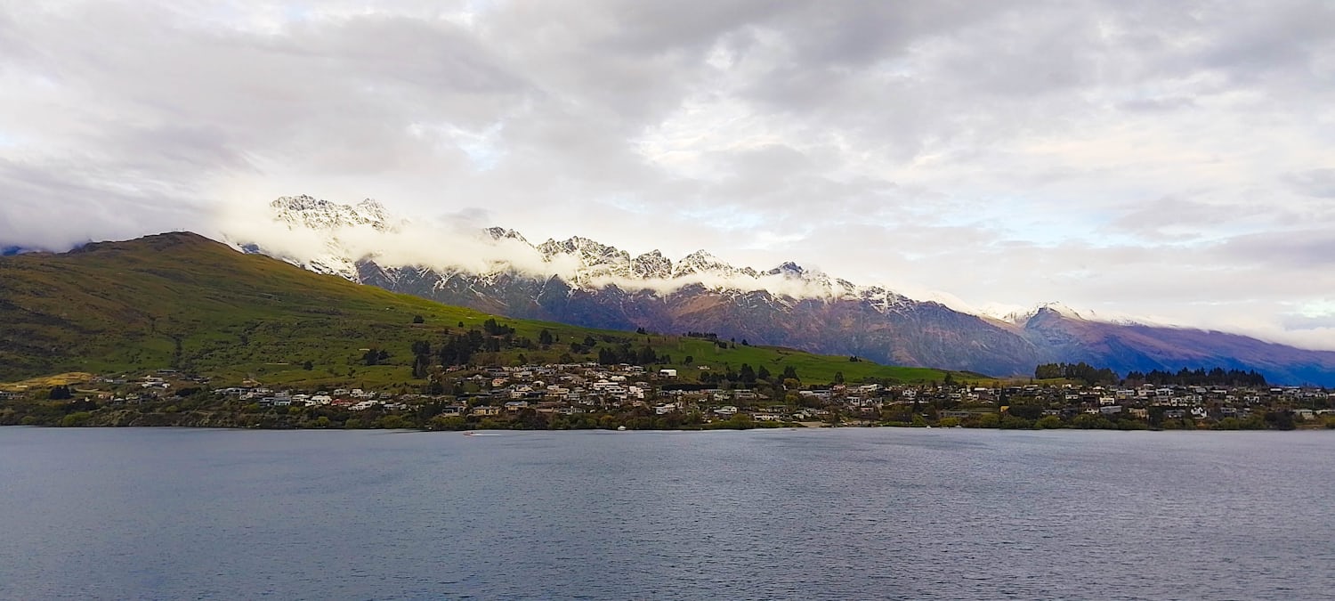 A wide shot of Queenstown across a lake. The sky is cloudy, and behind the town are mountains with snow-capped peaks.