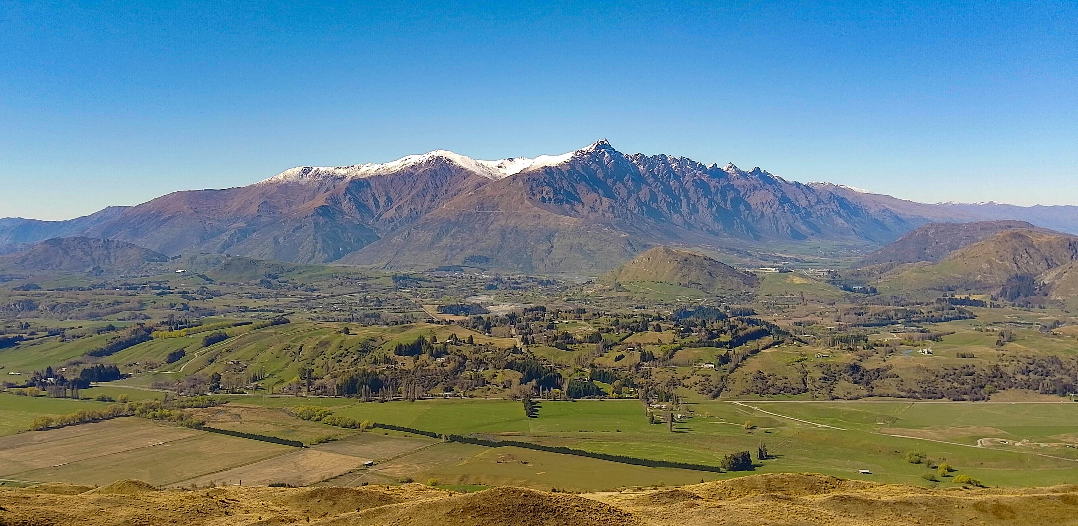 A wideshot of snow-capped mountains, with a valley sparsely populated with houses at the base. The sky is blue and clear.