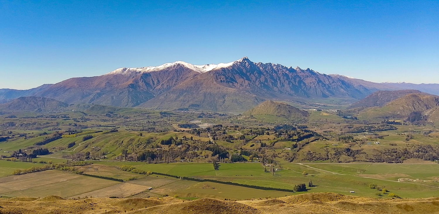 A wideshot of snow-capped mountains, with a valley sparsely populated with houses at the base. The sky is blue and clear.
