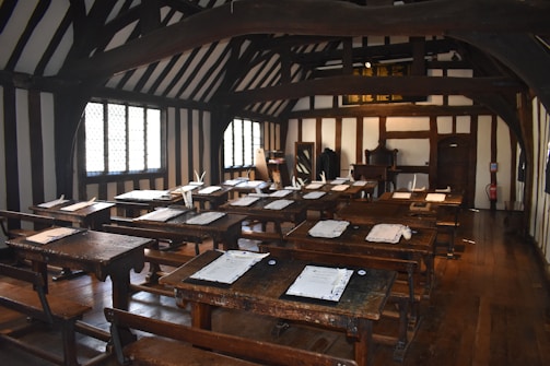 A historical classroom with wooden desks and benches arranged in rows. The walls and ceiling feature dark wooden beams contrasted against the white plaster, creating a Tudor-style interior. Sheets of paper with quills are placed on the desks, and sunlight streams through the lattice windows.