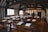 A historical classroom with wooden desks and benches arranged in rows. The walls and ceiling feature dark wooden beams contrasted against the white plaster, creating a Tudor-style interior. Sheets of paper with quills are placed on the desks, and sunlight streams through the lattice windows.