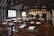A historical classroom with wooden desks and benches arranged in rows. The walls and ceiling feature dark wooden beams contrasted against the white plaster, creating a Tudor-style interior. Sheets of paper with quills are placed on the desks, and sunlight streams through the lattice windows.