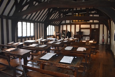A historical classroom with wooden desks and benches arranged in rows. The walls and ceiling feature dark wooden beams contrasted against the white plaster, creating a Tudor-style interior. Sheets of paper with quills are placed on the desks, and sunlight streams through the lattice windows.