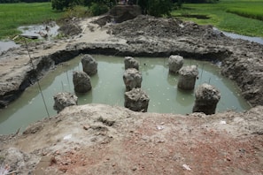 A construction site featuring several concrete pillars protruding from a muddy, water-filled pit. The surrounding area is marked by piles of dirt and rough terrain, with some greenery visible in the background.