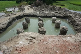 A construction site featuring several concrete pillars protruding from a muddy, water-filled pit. The surrounding area is marked by piles of dirt and rough terrain, with some greenery visible in the background.