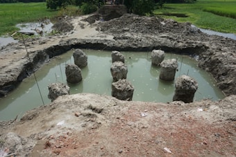 A construction site featuring several concrete pillars protruding from a muddy, water-filled pit. The surrounding area is marked by piles of dirt and rough terrain, with some greenery visible in the background.