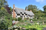 A charming thatched cottage with half-timbered walls is surrounded by lush greenery and a well-tended garden. Several people are engaged in gardening activities, hinting at a lively and productive atmosphere.