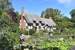A charming thatched cottage with half-timbered walls is surrounded by lush greenery and a well-tended garden. Several people are engaged in gardening activities, hinting at a lively and productive atmosphere.