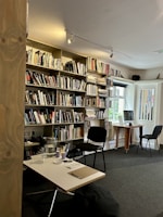 A cozy library or study room with a large bookshelf filled with various books. There are two chairs, one in front of a computer on a wooden desk and another near a smaller table with a notepad and pen. The space is well-lit with natural light coming from a window.