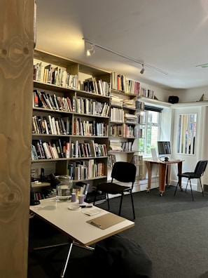 A cozy library or study room with a large bookshelf filled with various books. There are two chairs, one in front of a computer on a wooden desk and another near a smaller table with a notepad and pen. The space is well-lit with natural light coming from a window.