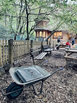 A wooded outdoor area with a dirt path featuring a wheelbarrow resting next to benches and a wooden fence. Background includes a rustic building with warm lighting, suggesting a cozy gathering space where people are sitting around a table.