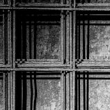 An intricate pattern of metal rebar with visible texture, forming a grid-like structure. The play of light and shadow creates a dynamic, geometric composition on a rough background.