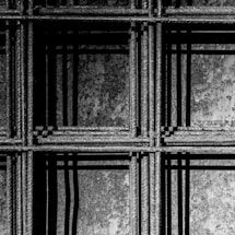 An intricate pattern of metal rebar with visible texture, forming a grid-like structure. The play of light and shadow creates a dynamic, geometric composition on a rough background.