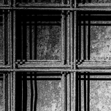 An intricate pattern of metal rebar with visible texture, forming a grid-like structure. The play of light and shadow creates a dynamic, geometric composition on a rough background.
