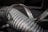 A close-up of a metallic coil with spiraling loops and visible texture, showing weathering and signs of wear. The focus highlights the ridged pattern and sheen of the metal under soft lighting, while the background is blurred.