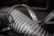 A close-up of a metallic coil with spiraling loops and visible texture, showing weathering and signs of wear. The focus highlights the ridged pattern and sheen of the metal under soft lighting, while the background is blurred.