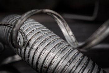 A close-up of a metallic coil with spiraling loops and visible texture, showing weathering and signs of wear. The focus highlights the ridged pattern and sheen of the metal under soft lighting, while the background is blurred.