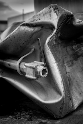 A close-up view of a crumpled and dented metal container with a pipe fitting protruding from one side. The surface of the metal appears weathered and textured with some dark stains and patches. The overall image is in black and white, adding a dramatic and gritty feel.