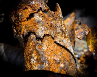 A close-up of a heavily rusted and decaying piece of metal. The surface is flaking and peeling, revealing a mix of orange, brown, and dark gray hues. The textures are rough and jagged, highlighting the extent of corrosion and oxidation.