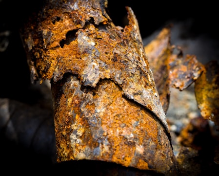 A close-up of a heavily rusted and decaying piece of metal. The surface is flaking and peeling, revealing a mix of orange, brown, and dark gray hues. The textures are rough and jagged, highlighting the extent of corrosion and oxidation.