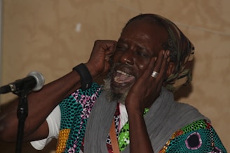 A person wearing colorful patterned clothing and a headscarf is passionately singing or speaking into a microphone. They have their eyes closed and hands placed near their ears, conveying a sense of deep engagement with the performance.