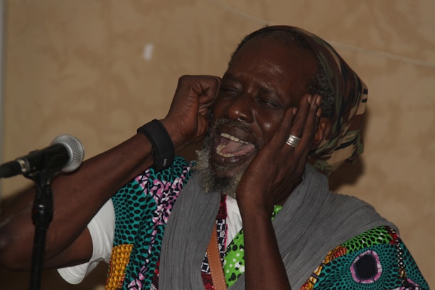 A person wearing colorful patterned clothing and a headscarf is passionately singing or speaking into a microphone. They have their eyes closed and hands placed near their ears, conveying a sense of deep engagement with the performance.
