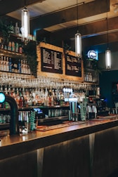 A dimly lit bar with a long counter, various bottles on shelves, hanging wine glasses, and illuminated pendant lights. The décor includes a chalkboard-style menu and beer taps glowing in blue light.